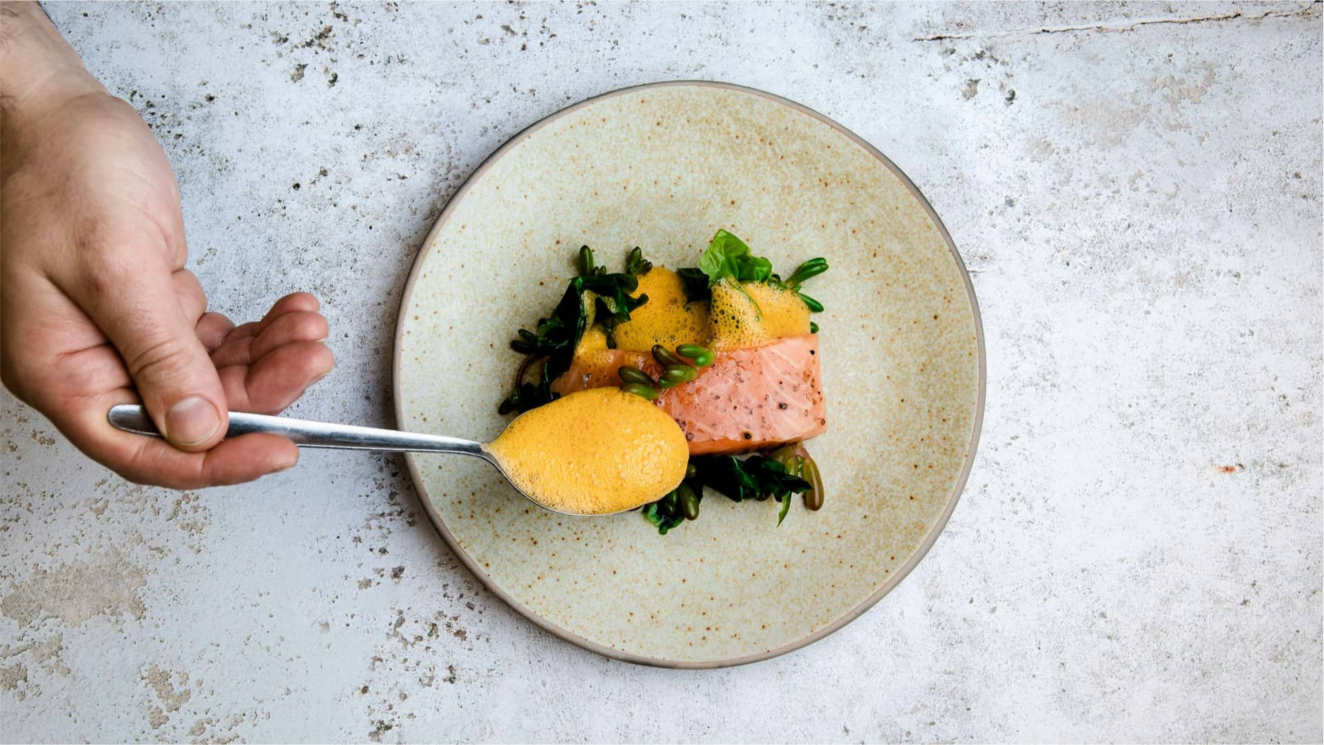 Above view of a man's hand holding a spoon and tipping orange sauce onto a plate of cooked salmon and vegetables