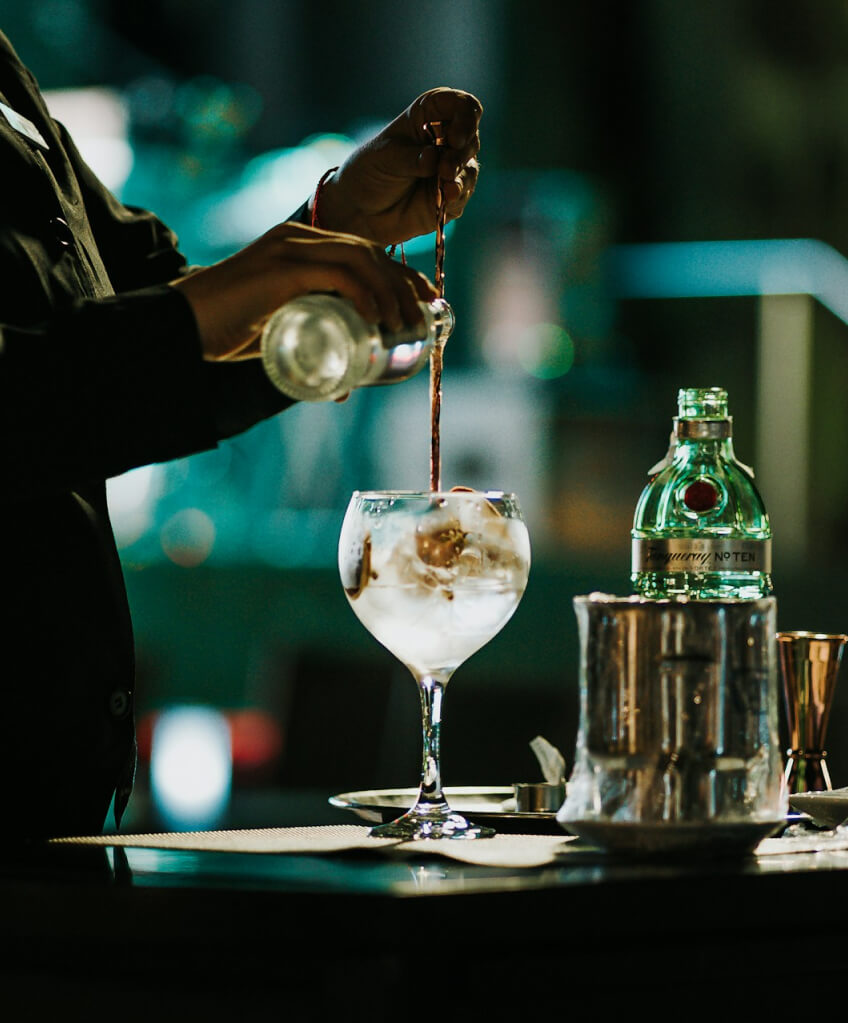 A distant side shot of a bartender pouring a clear liquor into a glass and stirring it.