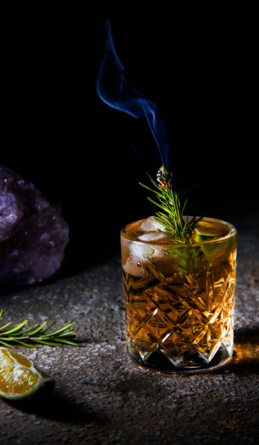 A front shot of a glass of whisky with a smoking rosemary sprig and ice inside it. Next to the glass is a slice of lime and a romseary sprig.